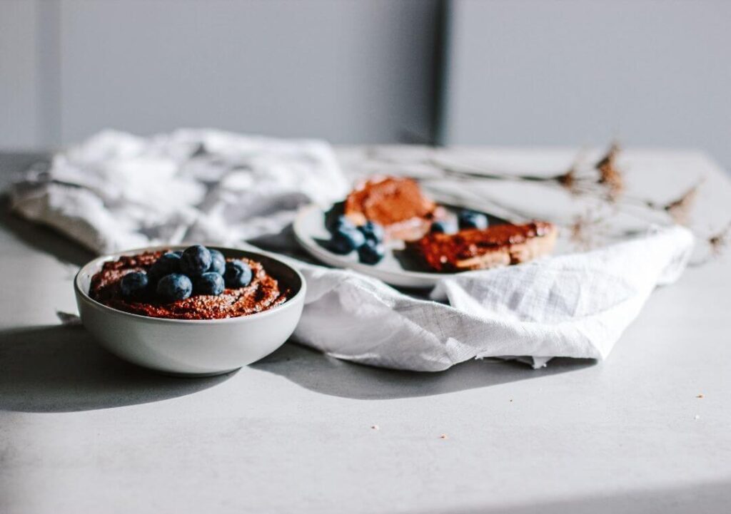 Schüssel mit veganer Schoko Hummus, Blaubeeren- Teller im Hintergrund bestrichen mit Schoko Hummus